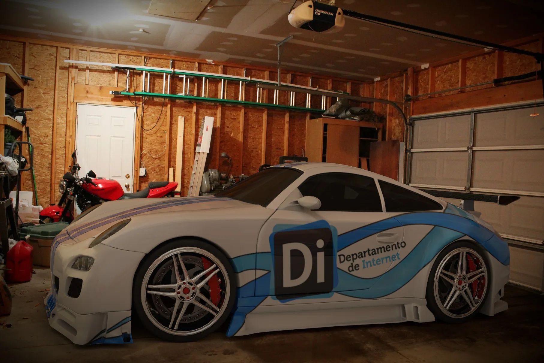 A white sports car with blue swooshes and a “Di Departamento de Internet” livery parked in a cluttered wooden garage, with a red motorbike behind it.